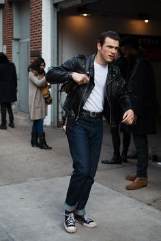 a man in black jacket and white t - shirt standing on sidewalk next to people