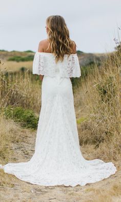 a woman in a white wedding dress standing on a dirt road looking off into the distance