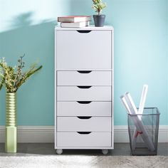 a tall white cabinet with five drawers next to a vase and plant on the floor