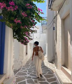 a woman walking down an alley way with pink flowers on the trees above her head