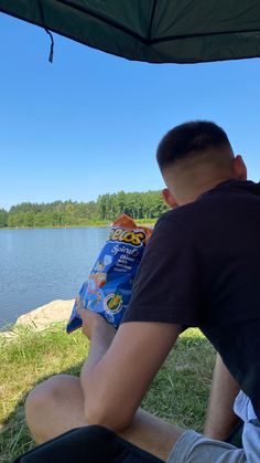 a man sitting on the ground next to a lake with an umbrella over his head