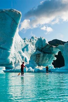 two people stand on surfboards in the water near icebergs