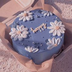 a blue birthday cake with white flowers on it sitting in a cardboard box next to some doily