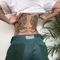 a person with tattoos on their back standing in front of a potted plant and wearing green pants