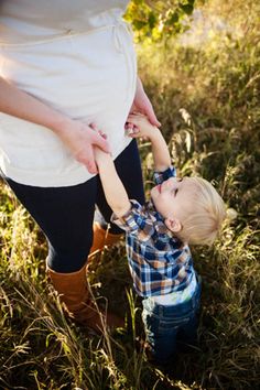 a woman holding the hand of a little boy in a field with tall grass and trees