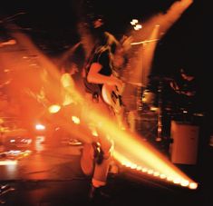 a man playing guitar on stage with other people in the background and lights behind him