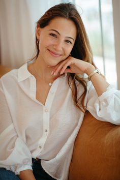 a woman sitting on a couch smiling at the camera with her hand under her chin