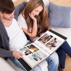 a man and woman sitting on a couch looking at an open photo album with pictures