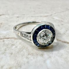 Breathtaking handcrafted platinum Art Deco style diamond and sapphire halo engagement ring. The center stone is a lovely bezel-set 0.50 carat old European cut diamond. The stone is G-H color VS2 clarity. It is haloed by a row of fine French cut sapphires weighing approximately 0.45 carat. The ring is accented with 4 round diamonds on the shoulders, weighing approximately 0.04 carat. G-H VS. Fine milgrain adds to the delicacy of the ring. Beautiful openwork filigree decorate the under gallery. Di Sapphire Diamond Ring With Rose Cut Diamonds, Lab-created Sapphire Diamond Ring With Halo Design, Vintage Round Diamond Ring With Halo, Diamond White Sapphire Ring With Rose Cut Diamonds, Round Sapphire Diamond Ring With Bezel Setting, Heirloom Sapphire Ring With Halo Design, Heirloom Sapphire Halo Ring, Vintage Round Halo Diamond Ring, Art Deco Diamond Ring With Bezel Setting