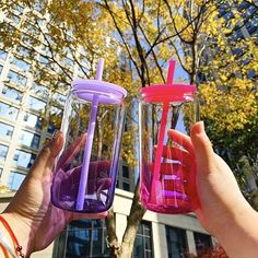 two people are holding up colorful cups in front of a tall building with trees and buildings behind them