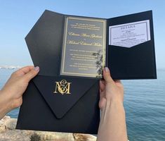 a person holding up an open black and gold wedding card with the ocean in the background