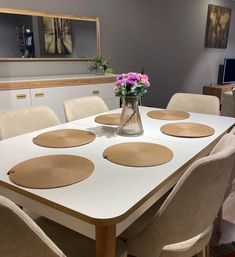 a vase with flowers on top of a white table surrounded by beige chairs and placemats