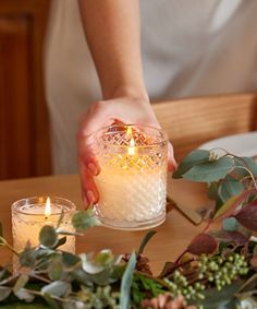 a person holding a lit candle on top of a table next to flowers and greenery