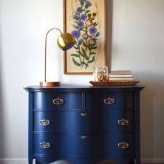 a blue dresser with flowers on it and a painting hanging above the top, next to a lamp