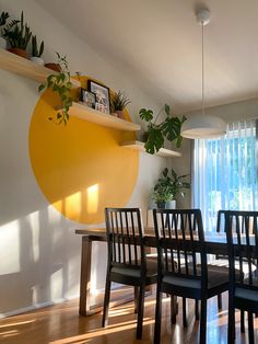 a dining room table and chairs with plants on the shelf above them in front of a window