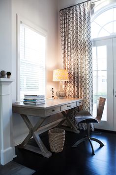 a room with a desk, chair and lamp in it next to a window covered by curtains