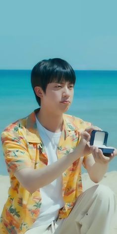 a young man sitting on the beach looking at his cell phone while holding something in his hand