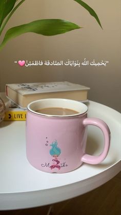 a pink coffee cup sitting on top of a white table next to a green plant