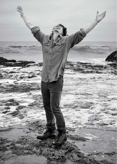 a man standing on top of a beach next to the ocean with his hands in the air