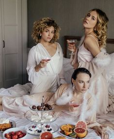 two women in white dresses sitting on a bed with fruit and pastries around them