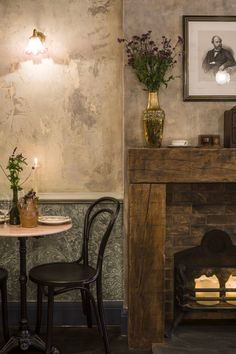 two chairs and a table in front of a fireplace with flowers on the mantel