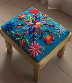 a small wooden stool with colorful embroidered flowers on the top and bottom, sitting in front of a window