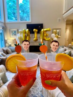 two people holding up drinks with orange slices and straws on them in front of a living room