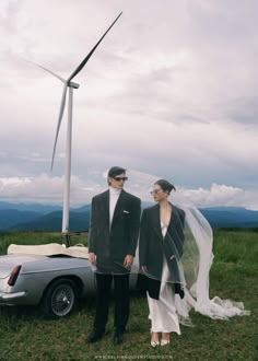 a bride and groom standing in front of a car with a wind turbine behind them