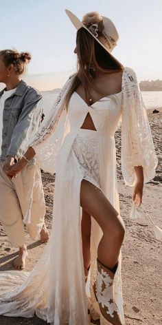 a woman in a white dress and cowboy boots standing on the beach next to another woman