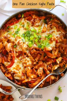 a skillet filled with pasta and vegetables on top of a white table next to silver spoons