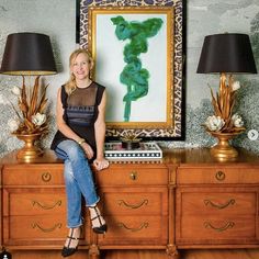 a woman sitting on top of a dresser in front of a painting and two lamps