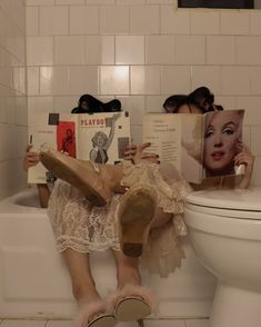 a woman sitting on a toilet reading a book with her feet up in the bathtub