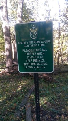 a green sign that is in the middle of some grass and dirt with trees behind it