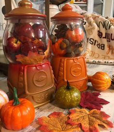 two glass jars filled with different types of fall leaves and pumpkins on a table