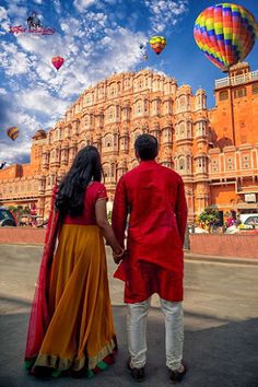 a man and woman standing in front of a building with hot air balloons flying overhead