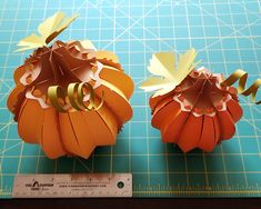 two paper pumpkins sitting on top of a cutting board next to a measuring ruler