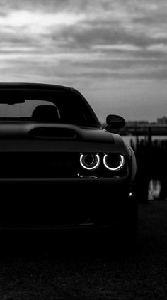 a black and white photo of a car with the headlights turned on, in front of a body of water