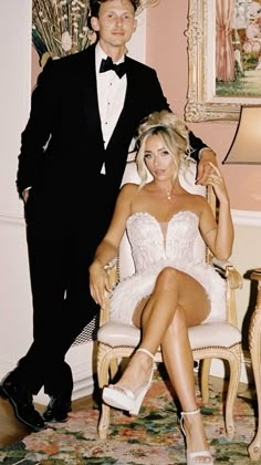 a man in a tuxedo standing next to a woman sitting on a chair