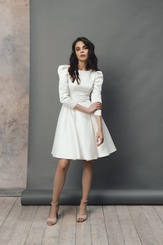 a woman standing in front of a gray wall wearing a white dress with ruffle sleeves