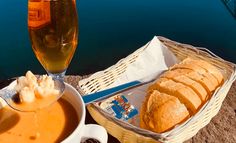 a basket of bread and soup next to a glass of beer on a table by the water