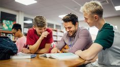 three young men sitting at a table in a library, one reading a book and the other smiling