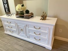 a white dresser with gold handles and flowers on top is in the corner of a room