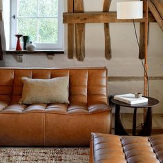 a brown leather couch sitting in front of a window next to a table with a lamp on it