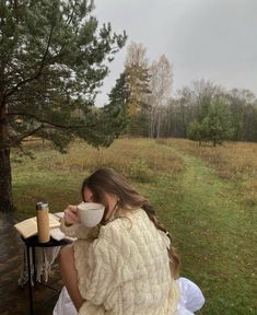 a woman sitting at a picnic table drinking from a coffee cup while wearing a knitted shawl