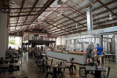 the inside of a building with tables, chairs and people standing around it in front of a bar