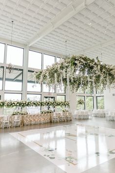 an indoor wedding venue with white chairs and floral arrangements hanging from the ceiling, surrounded by large windows