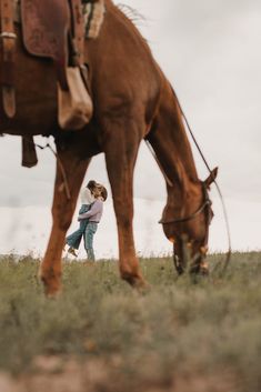 a woman walking next to a brown horse on top of a grass covered field with a sky background