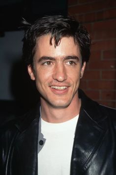 a man wearing a black leather jacket and white t - shirt smiles at the camera