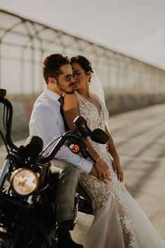 a bride and groom are sitting on a motorcycle in front of a bridge with their arms around each other