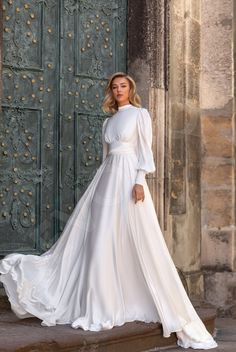 a woman standing in front of a door wearing a white dress with long sleeves and a bow at the waist